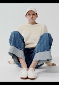 Young person wearing a cream sweatshirt, wide-cuffed blue jeans, white cap, and white shoes, sitting on the floor against a plain background.