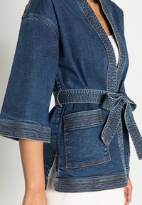 Jeansowy płaszcz w stylu kimona w ciemnoniebieskim kolorze, z szerokimi rękawami, zaciągniętym paskiem w talii oraz dwiema przednimi kieszeniami z kontrastującymi detalami przeszyć.