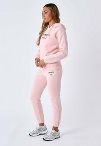 Felpa con cappuccio rosa chiaro con testo "PROJECT X PARIS", abbinata a pantaloni sportivi rosa coordinati. Tessuto testurizzato, polsini elasticizzati e design sportivo.