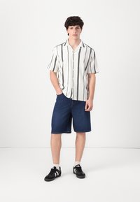 Chemise blanche à manches courtes avec des rayures verticales noires, assortie à un short en denim bleu et des baskets noires avec des accents blancs.