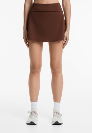 Brauner sportlicher Rock mit flachem Bund und ausgestelltem Design. Kombiniert mit weißen Knöchelsocken und hellen Sneakern.