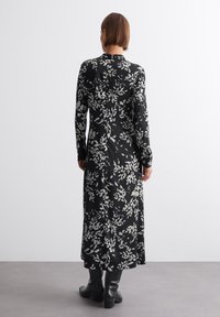 Langermet svart kjole med hvitt blomsterdesign, med en tettsittende kropp og en flytende skjørt. Laget av lett stoff.