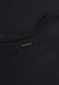 Zbliżenie na czarny materiał, ukazujące obszar kołnierza z małą metalową metką z napisem "SUPERDRY" przymocowaną poniżej dekoltu.