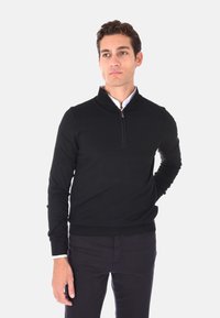 Maglione nero a mezze zip realizzato in un tessuto a maglia liscia, presenta un orlo e polsini a costine, abbinato a pantaloni scuri e una camicia con colletto sotto.