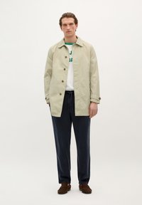 Trench coat bej deschis cu nasturi maro, purtat peste o cămașă albă cu text verde, asortat cu pantaloni bleumarin închisi și mocasini maro.