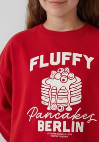 Rode sweatshirt met witte grafische tekst "FLUFFY" en een illustratie van gestapelde pannenkoeken met toppings, met de detailtekst "Pannenkoeken BERLIJN."
