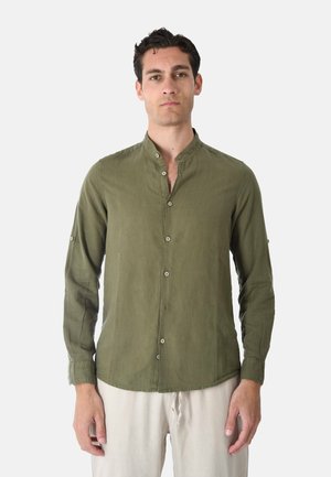 Uomo che indossa una camicia verde oliva a manica lunga con colletto alla mandarino e pantaloni beige chiaro, in piedi davanti a uno sfondo neutro.