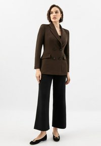 Blazer doppiopetto marrone con bottoni neri, due tasche frontali, abbinato a pantaloni neri a gamba larga e décolleté neri con dettagli dorati.