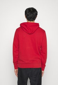 Homme aux cheveux noirs courts portant un sweat à capuche rouge et un pantalon noir, debout face à un fond blanc uni.