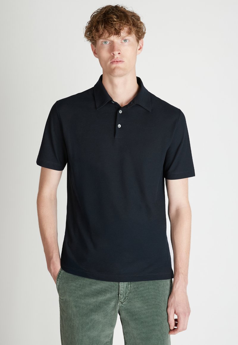Camisa polo negra con cuello clásico, mangas cortas, tapeta de tres botones y una textura suave de algodón. Llevada con pantalones de pana verdes.