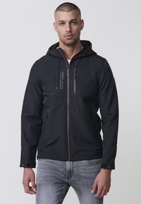 Jacheta neagră din softshell are un fermoar frontal, glugă, două buzunare laterale și accente de cusătură contrastante. Textură netedă, material ușor.