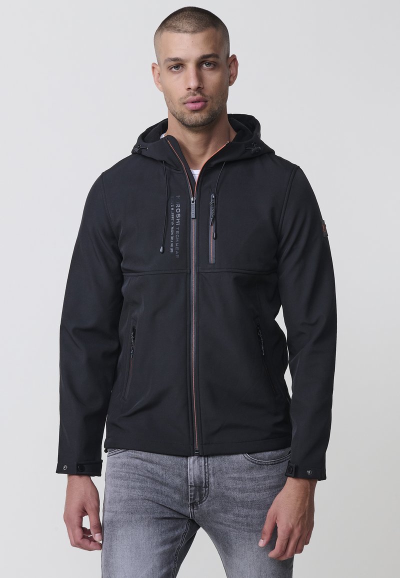 Jacheta neagră din softshell are un fermoar frontal, glugă, două buzunare laterale și accente de cusătură contrastante. Textură netedă, material ușor.