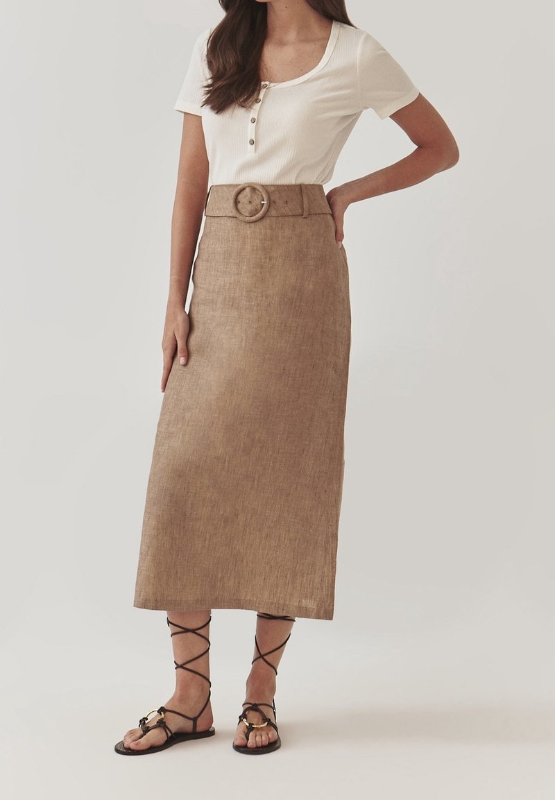 Leniowa maxi spódnica w jasnobrązowym kolorze z szerokim paskiem i okrągłą klamrą, zestawiona z białą koszulą na krótki rękaw zapinaną na guziki oraz czarnymi płaskimi sandałami.