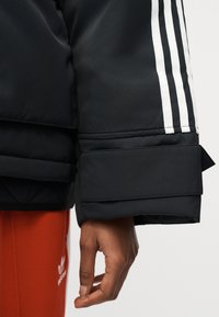 Κοντινό πλάνο ενός χεριού που κρέμεται στο πλάι, φορόντας μαύρο μπουφάν με λευκές ρίγες και κόκκινο παντελόνι Adidas με λογότυπο.