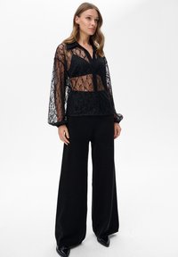 Czarna, przezroczysta bluzka z koronką, zapinana na guziki, z kołnierzykiem i długimi rękawami, noszona na czarnym topie; zestawiona z szerokimi, czarnymi spodniami z wysokim stanem.