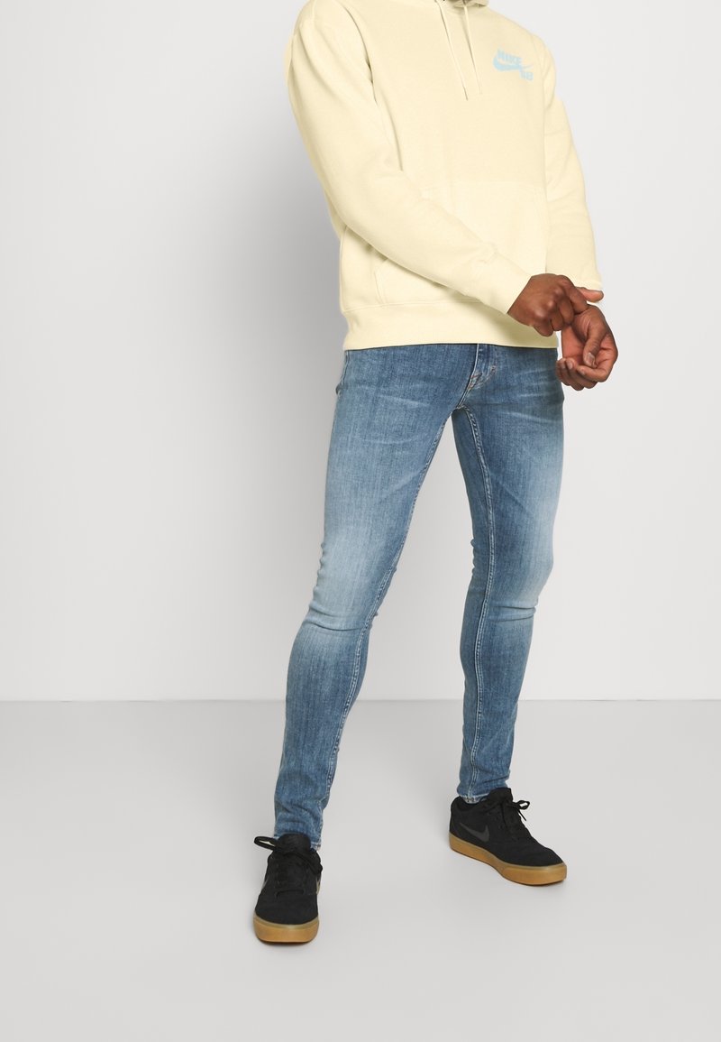 Personne portant un sweat à capuche beige clair, un jean skinny bleu clair et des baskets noires à semelles en gomme, se tenant devant un fond uni clair.