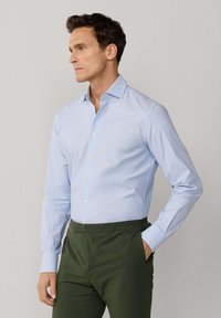 Homme portant une chemise à boutons bleu clair et un pantalon vert foncé, debout avec une main dans la poche, regardant vers la gauche sur un fond uni.