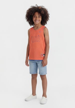 Enfant souriant aux cheveux bouclés portant un haut sans manches orange Nike, un short en jean bleu clair et des chaussures blanches sans lacets, debout sur fond blanc.