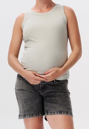 Zwangere persoon draagt een licht mouwloos topje en donkere denimshorts, houdt zachtjes haar buik met beide handen vast.
