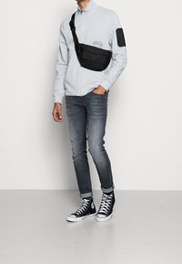 Person som bär en ljusgrå zip-up jacka med en svart korsbody-väska, urblekta grå jeans och svarta högstövlar med vita snören.