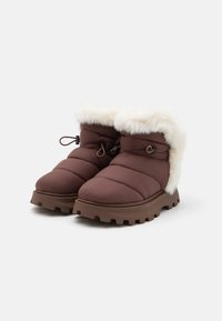 Brune polstrede støvler med en hvid falsk pelskant, der har en gummisål med et robust mønster og dekorative snørebånd for justerbar pasform.