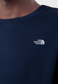 Gros plan d'une personne portant une chemise bleu foncé avec un petit logo blanc The North Face sur le côté gauche de la poitrine.