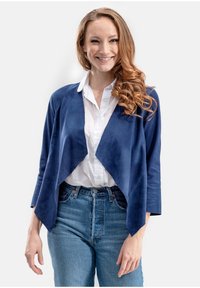 Marineblauwe suède drapeerjas met open voorkant, driekwartmouwen, gecombineerd met een witte blouse en hooggewase blauwe jeans.