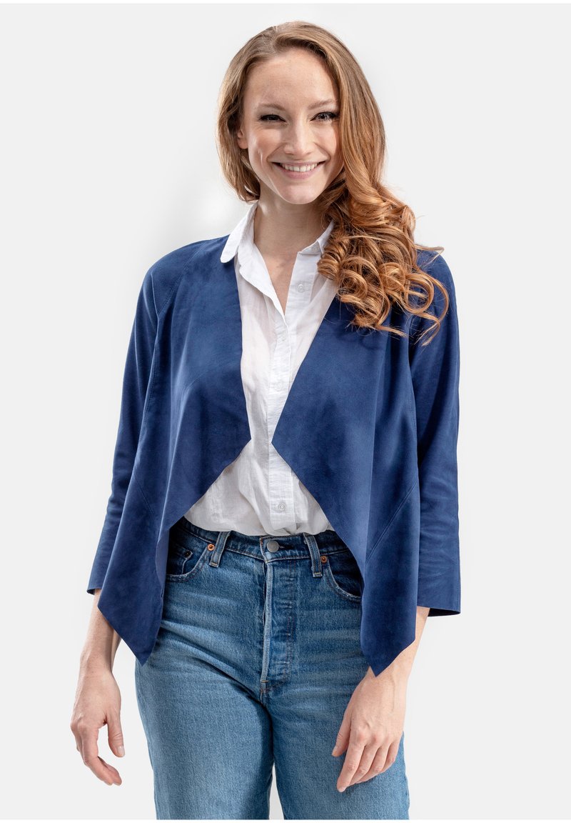 Marineblauwe suède drapeerjas met open voorkant, driekwartmouwen, gecombineerd met een witte blouse en hooggewase blauwe jeans.
