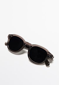 Tmavě tónované nadměrné sluneční brýle s průhledným šedým rámem. Mají kulaté čočky, rovná ramínka a unikátní geometrický design.