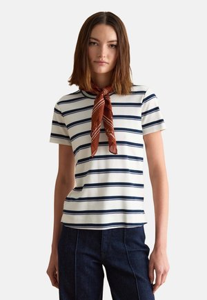 Jonge vrouw die een wit en marineblauw gestreept shirt met korte mouwen draagt, donkere spijkerbroek en een gebonden bruine sjaal met patroon om haar nek, staande tegen een effen achtergrond.