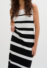 Donna in un vestito senza maniche a righe orizzontali nere e bianche in piedi contro uno sfondo neutro, mano che stringe il tessuto del vestito vicino alla vita.