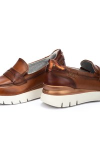 Zapatos de piel marrón tipo slip-on con un acento metálico, que cuentan con una suela blanca gruesa y tachuelas decorativas en la parte trasera. Textura suave en toda la superficie.