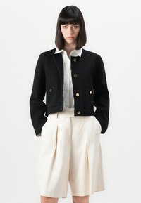 Veste noire courte avec des boutons dorés, deux poches avant et une texture lisse, associée à un short plissé crème et à une chemise blanche.