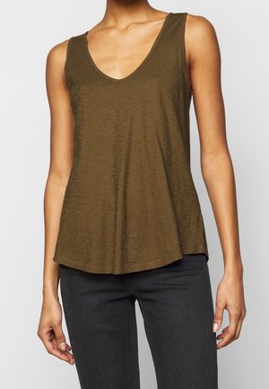 Vrouw die een olijfgroene mouwloze top met V-hals en donkergrijze nauwsluitende broek draagt, staand met ontspannen armen tegen een effen achtergrond.