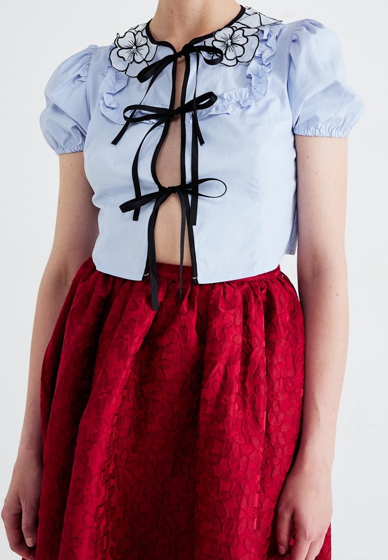 Blouse courte bleu clair avec manches bouffantes, liens rubans noirs, broderie florale au col, associée à une jupe rouge texturée à motif floral.