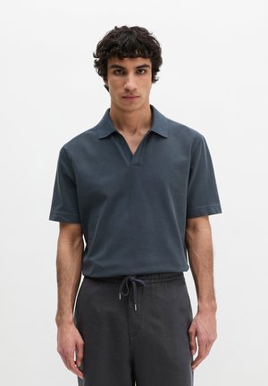 Jonge man met krullend haar, gekleed in een donkergrijs poloshirt met korte mouwen en donkere broek met trekkoord, staand tegen een effen witte achtergrond.