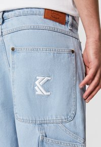 Ljusblå denimbyxor med en ficka med vit broderad logotyp och en brun läderlapp vid midjan, hand vilande bredvid fickan.