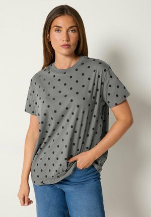 Femme portant un t-shirt gris à manches courtes avec des pois noirs et un jean bleu, debout, une main dans la poche, devant un fond uni.