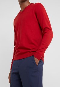 Czerwony sweter w serek o miękkiej fakturze, z długimi rękawami i ściągaczami, w połączeniu z granatowymi spodniami. Subtelny detal logo na klatce piersiowej.