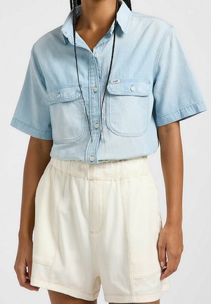Camicia di jeans azzurro chiaro a maniche corte con due tasche sul petto e bottoni bianchi, abbinata a shorts crema a vita alta con dettagli di cucitura.
