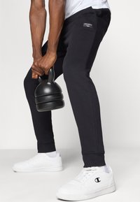 Calças de treino pretas com um design ajustado, com punhos nas pernas e bolsos laterais, combinadas com sapatilhas brancas e um kettlebell preto.