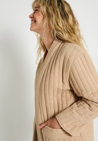 Femme blonde aux cheveux ondulés souriant, portant une veste matelassée beige à rayures verticales et une main dans la poche latérale, sur un fond uni.