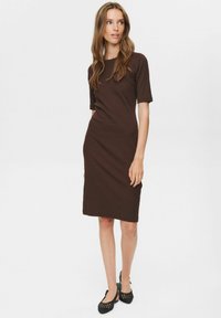 Kvinna som står i en brun knälång klänning med kort ärm och svarta vävda slingback-sandaler mot en enfärgad bakgrund.