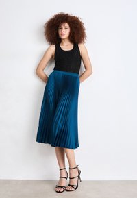 Top in paillettes nere lucenti abbinato a una gonna pieghettata teal lunghezza ginocchio. Tacchi neri con cinturini completano l'outfit. Sfondo neutro.