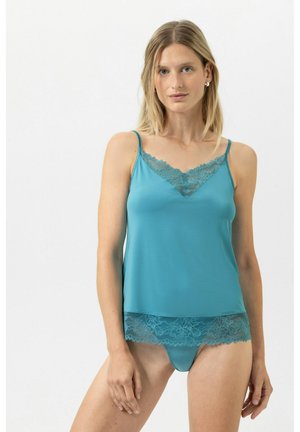 Vrouw met blond haar die een teal kanten hemdje en bijpassende slip draagt, staande tegen een effen witte achtergrond.