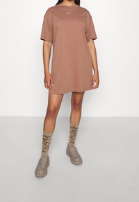 Brązowy oversize t-shirt z krótkim rękawem i niewielkim logo na piersi, w połączeniu z beżowymi butami typu slip-on i wzorzystymi beżowymi skarpetkami.