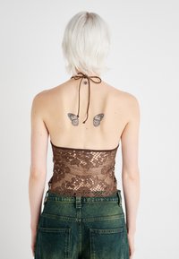 Hnedý halter top so zlato-poskladanými flitrami a otvoreným chrbtom, skombinovaný so zelenými džínsami s veľkými vreckami. Viditeľné sú dve tetovania motýľov.
