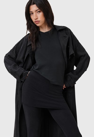 Trench-coat noir oversize porté sur un top noir ajusté et une jupe. Tissu lisse, manches longues, et coupe décontractée pour un look superposé.