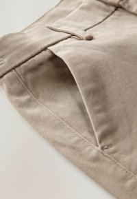 Pantaloni in cotone beige con una texture liscia, caratterizzati da un particolare tasca frontale e cuciture ordinate lungo i bordi.