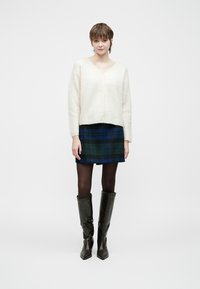 Kremfarget strikket genser med V-hals, kombinert med et rutet skjørt i mørkeblått og grønt. Kne-høye svarte skinnstøvler fullfører antrekket.